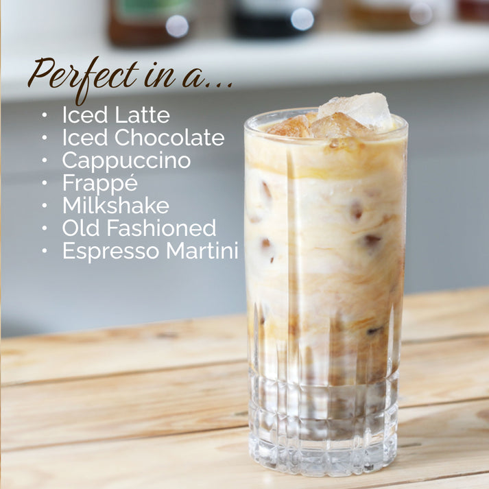 A tall glass filled with an iced latte sits on a wooden surface. Beside it, text reads: "Perfect in an... Iced Latte, Hot Chocolates, Cappuccino, Frappé, Milkshake, Old Fashioned, Espresso Martini." The flavor is completed with a hint of MONIN Hazelnut Syrup from the renowned brand MONIN. A blurry kitchen scene forms the background.